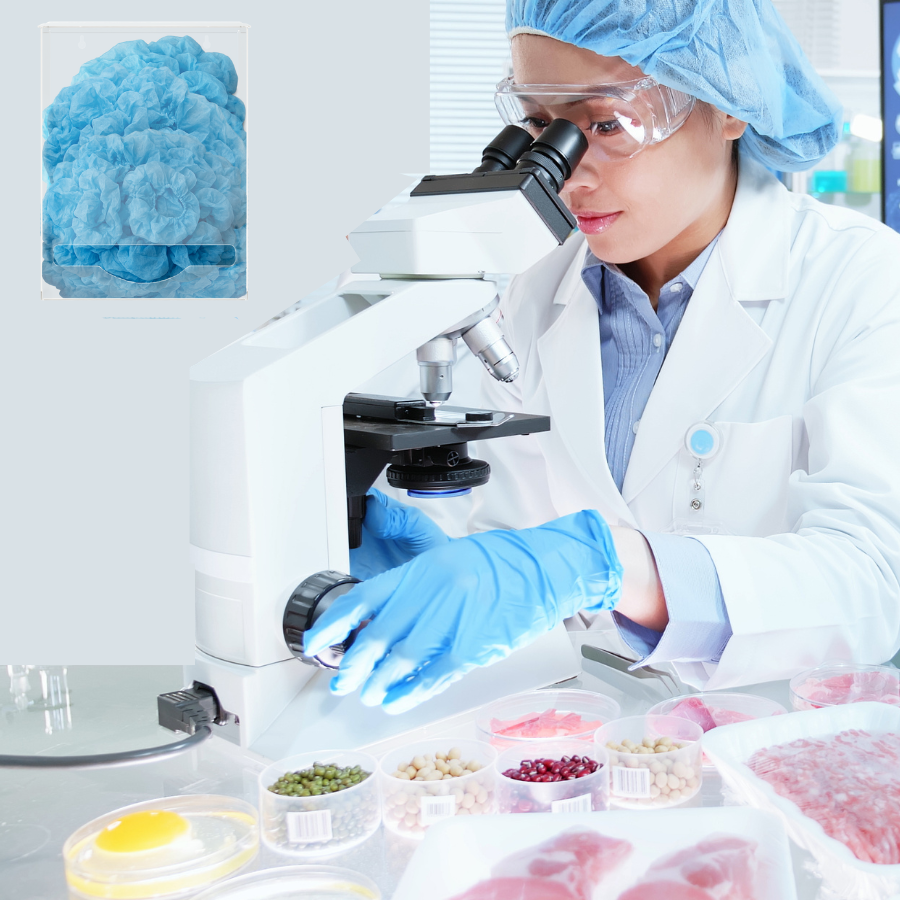 Lab worker in a food lab at a microscope with her PPE on goggles, bouffant cap, gloves looking into microscope with an ORIGIN single bulk dispenser on the wall to her side with bouffant caps inside.